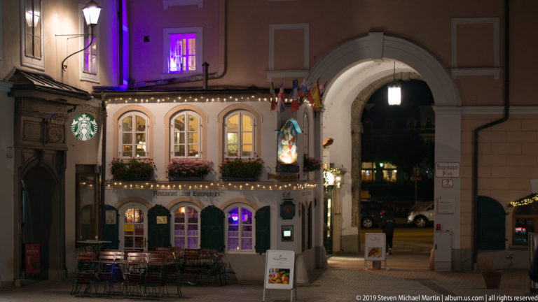 Illuminated cafe with outdoor tables beside a large rounded stone archway at night.
