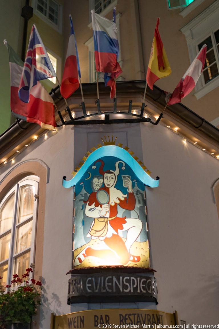 Painted Till Eulenspiegel jester sign beneath international flags on a Salzburg restaurant facade.