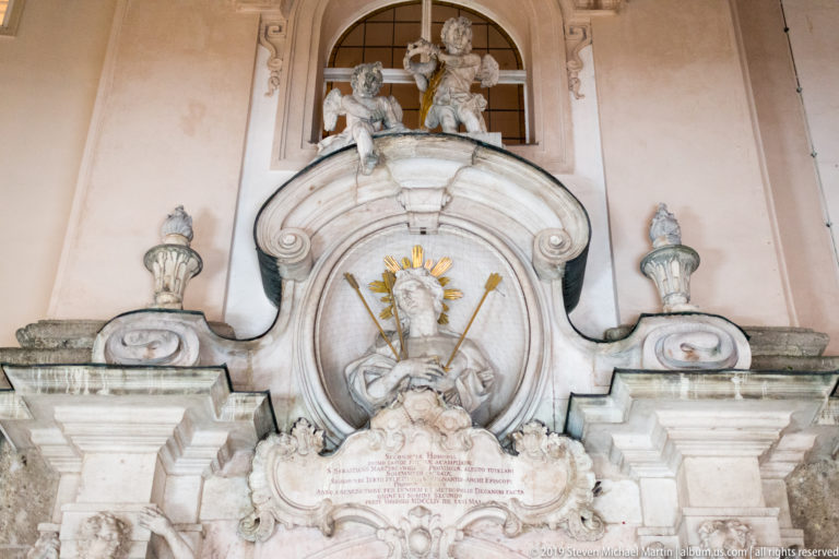 Ornate Baroque wall monument with portrait medallion, gilded rays, inscription, urns, scrollwork, and sculpted figures.
