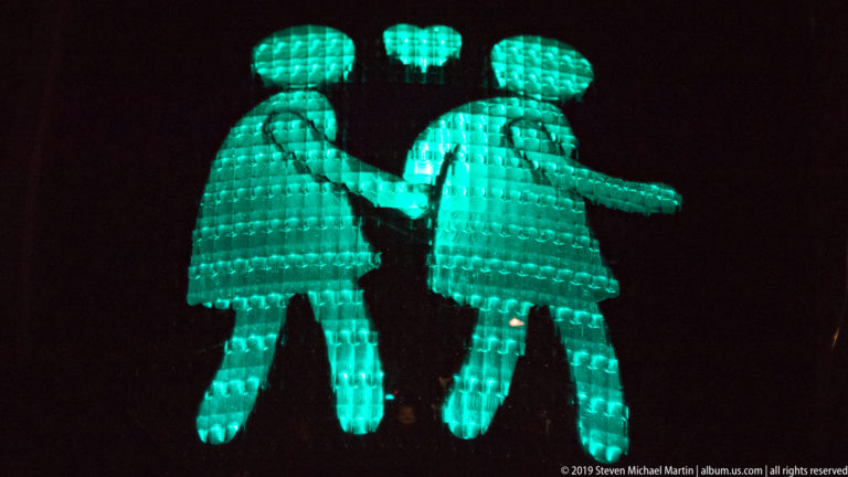 Two turquoise illuminated figures holding hands beneath a heart, with one figure wearing a skirt-like silhouette.
