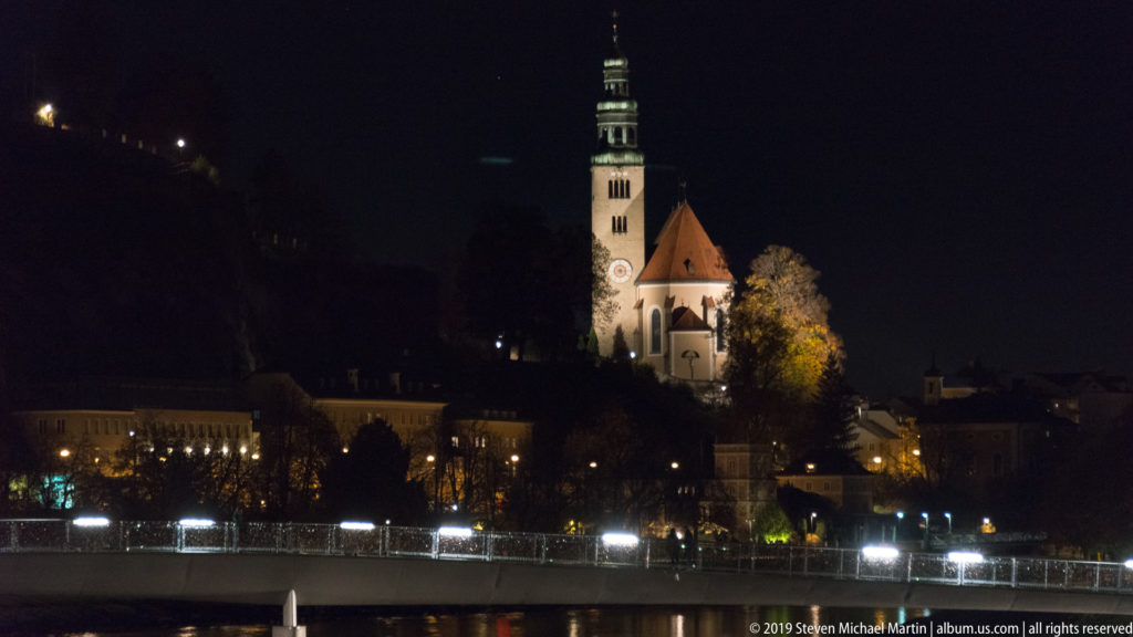 Illuminated hilltop church with clock tower and red roof above the Salzach River, with bright riverfront lights below.
