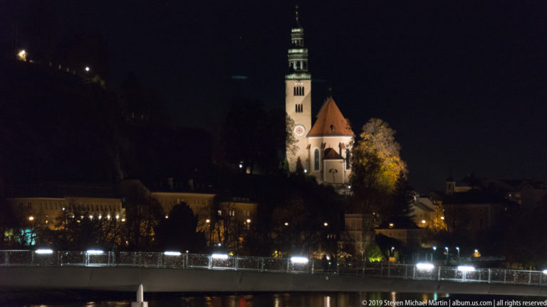 Illuminated church with tall clock tower and red roof above the Salzach River and bright waterfront lights.