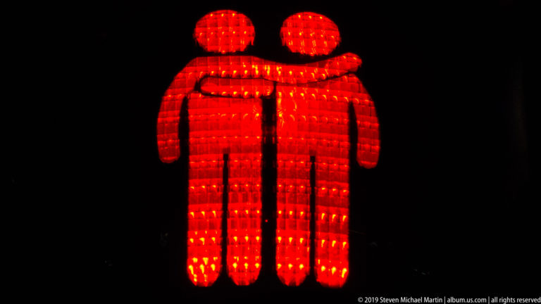 Red illuminated pictogram of two people standing together, one with an arm around the other's shoulders.
