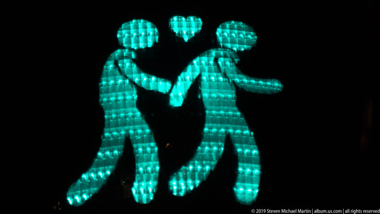 Turquoise illuminated figures holding hands beneath a small heart against a black nighttime background.