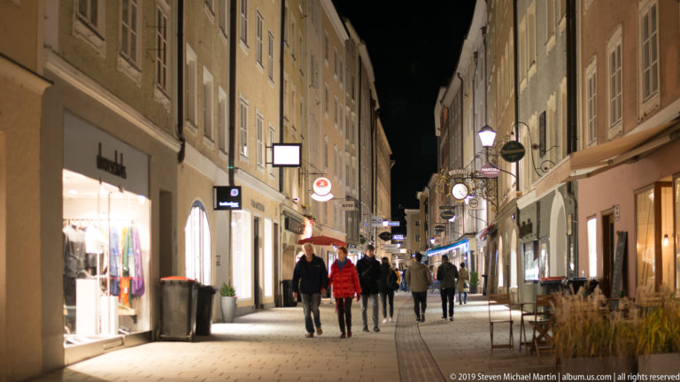 Narrow Salzburg street at night with illuminated shops, hanging signs, café furniture, and pedestrians walking away.