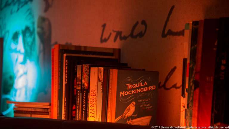 Cocktail books on a shelf under red and turquoise light, with Tequila Mockingbird facing the camera.