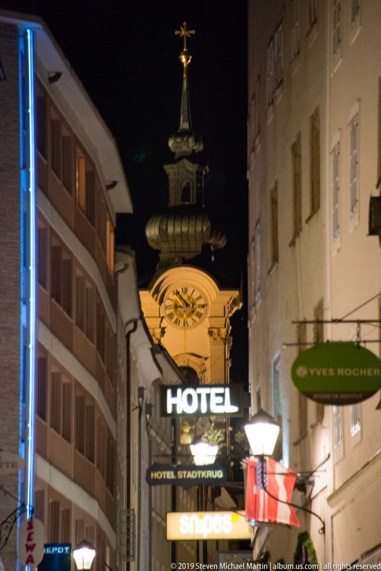 Night street with hotel and shop signs, an Austrian flag, and an illuminated clock tower rising behind.