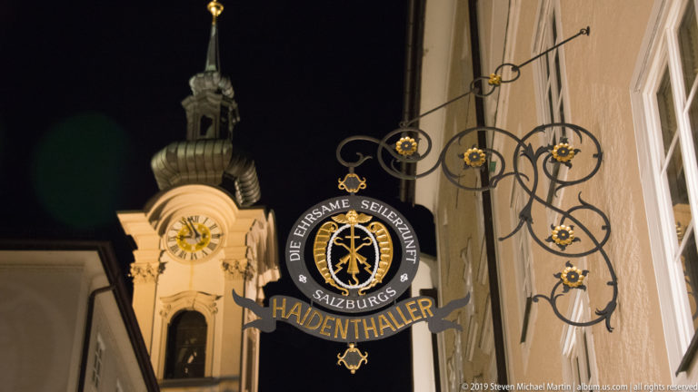 Ornate Haidenthaller hanging shop sign with gilded details silhouetted before an illuminated clock tower.