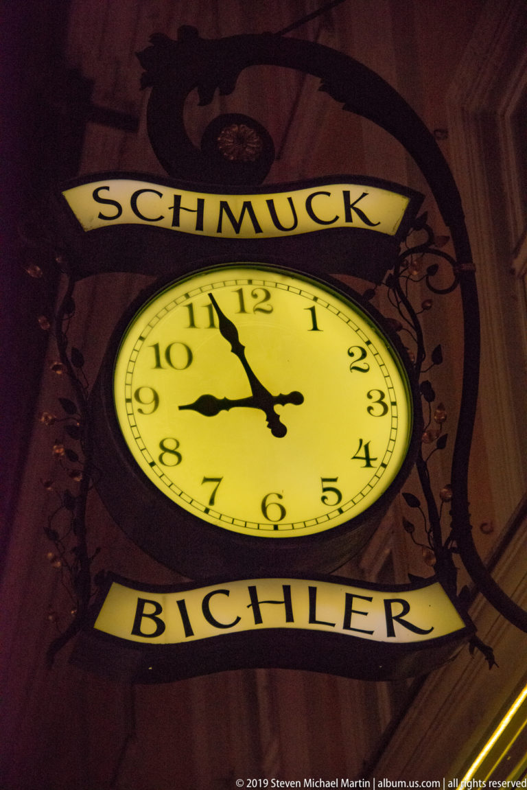 Illuminated yellow analog clock sign reading “SCHMUCK BICHLER” on a dark Salzburg street.
