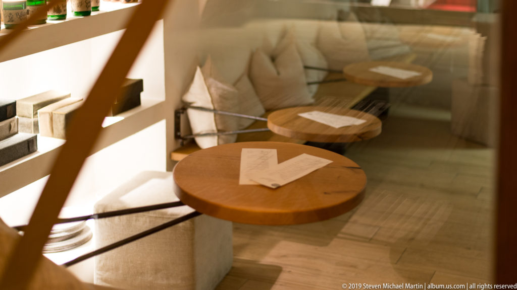 Round wooden cafe tables beside pale upholstered bench seating, viewed through a reflective window at night.