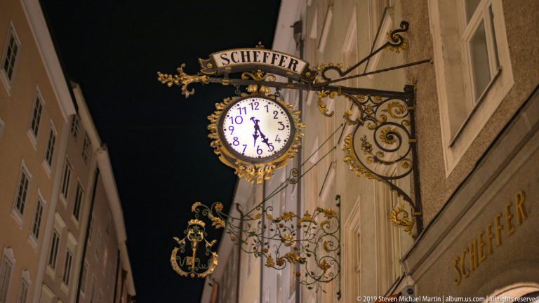 Ornate Scheffer projecting sign with illuminated clock, gilded scrollwork, and a smaller hanging crest at night.