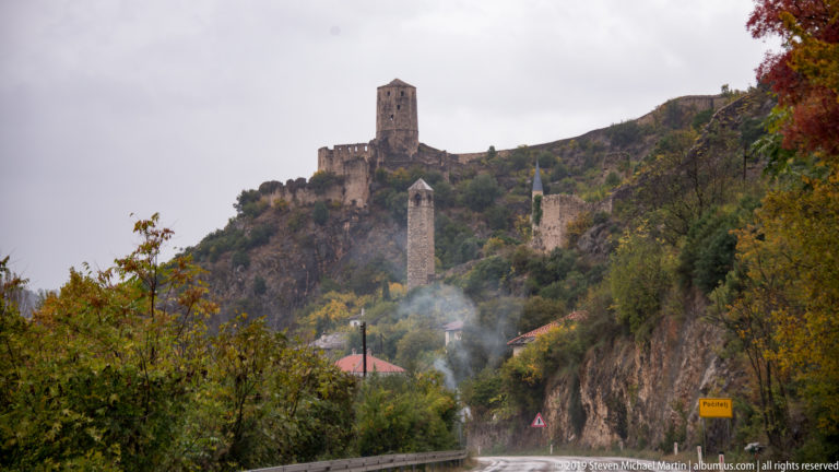 Počitelj fortress tower above a wooded hillside with lower stone towers, mosque minaret, smoke, and a curving road.