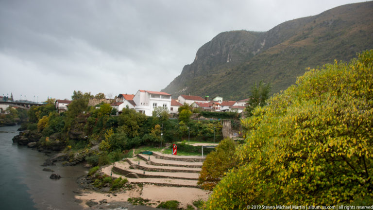 Neretva River beside stepped stone terraces, autumn trees, houses, and a steep mountain under cloudy skies.