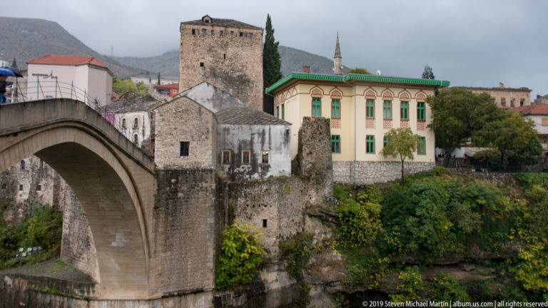 Stari Most arch beside a massive square stone tower and historic buildings above the Neretva River.