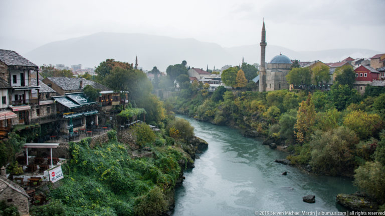 Neretva River running between rocky banks, riverside buildings, autumn trees, and a domed mosque with minaret.