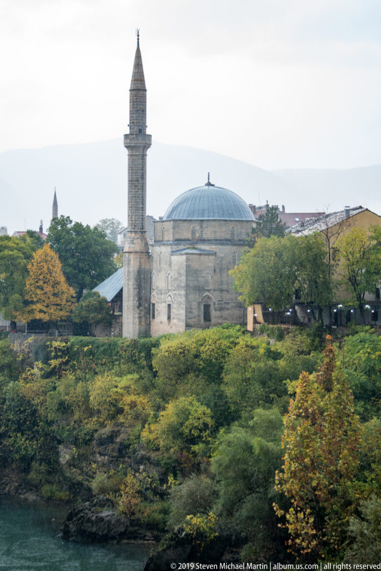 Side view of a stone mosque with a broad gray dome and tall minaret behind dense green and yellow vegetation.
