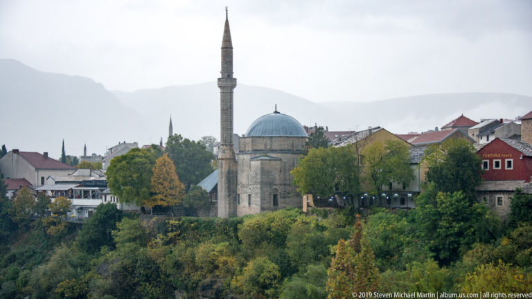 Domed stone mosque with a tall minaret rising above autumn trees, with hazy mountains in the background.