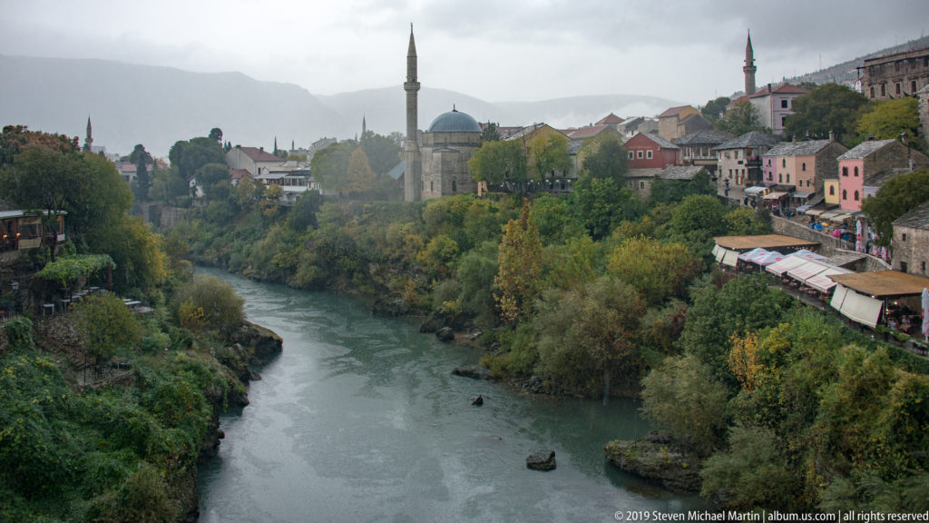 Neretva River flowing between wooded banks, with historic buildings, a domed mosque, and minarets above.