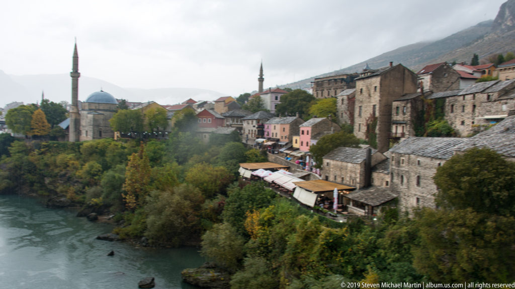 Neretva River curving past autumn trees, stone houses, colorful buildings, a domed mosque, and two minarets.