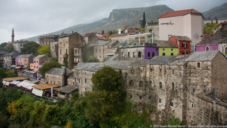 Large stone buildings and colorful houses on a steep riverbank in Mostar, with minarets and a mountain behind.
