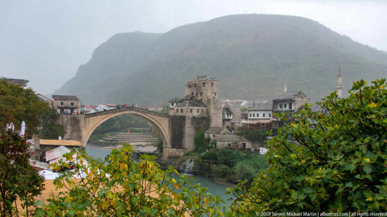 Stari Most spanning the Neretva River beneath a misty mountain, framed by autumn foliage and historic buildings.