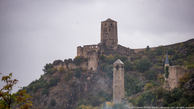 Tall stone fortress tower and crenellated walls above lower towers and a mosque minaret on Počitelj's hillside.