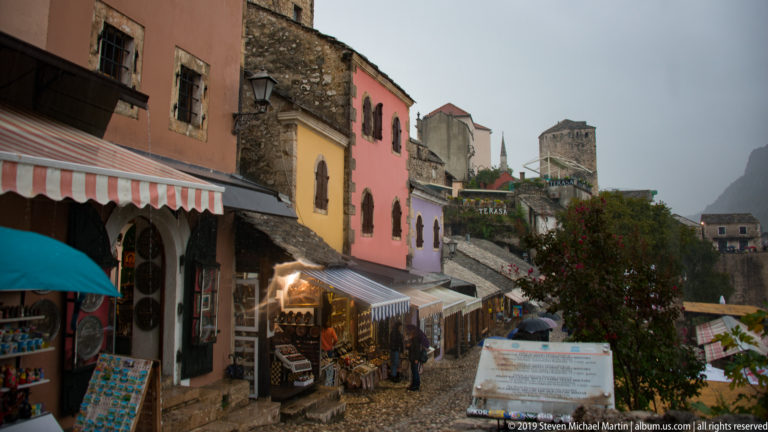 Cobbled market lane lined with awnings and colorful shopfronts, with stone buildings, a tower, and minaret beyond.