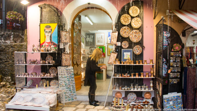 Shop entrance filled with decorated plates, jewelry, textiles, framed pictures, and small souvenirs under a stone arch.