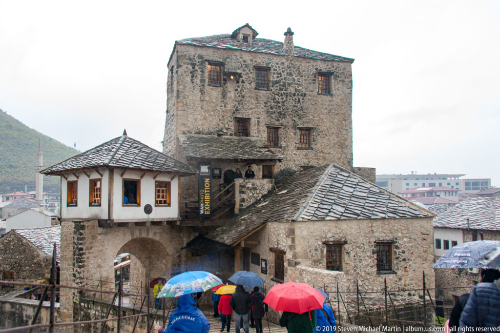 People with umbrellas approach an arched passage beneath tall historic stone buildings and gray roofs in Mostar.