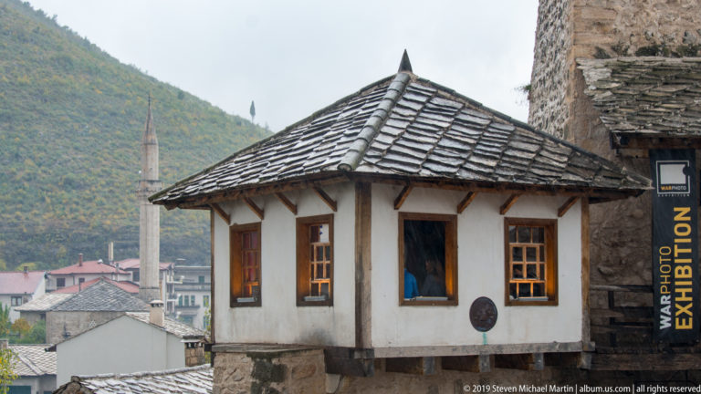 White traditional house with stone-slab hipped roof and mosque minaret rising behind it in Mostar.