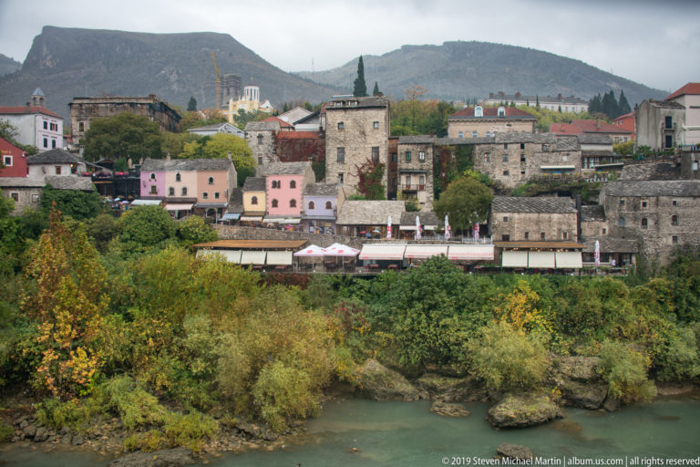Historic stone and pastel buildings across the Neretva River, backed by trees and a low mountain ridge.