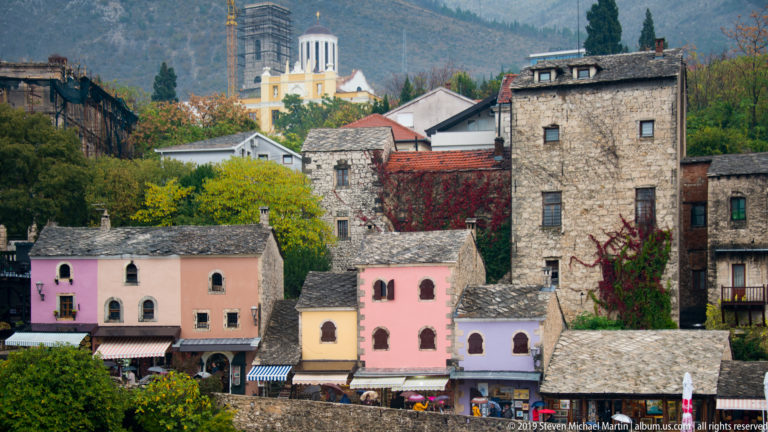 Pastel houses below tall stone buildings in Mostar, with autumn trees, red vines, and a pale church tower in the distance.