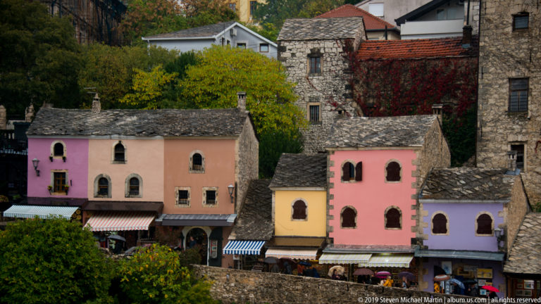 Pastel pink, yellow, and lavender houses with arched windows beneath taller stone buildings and gray roofs.