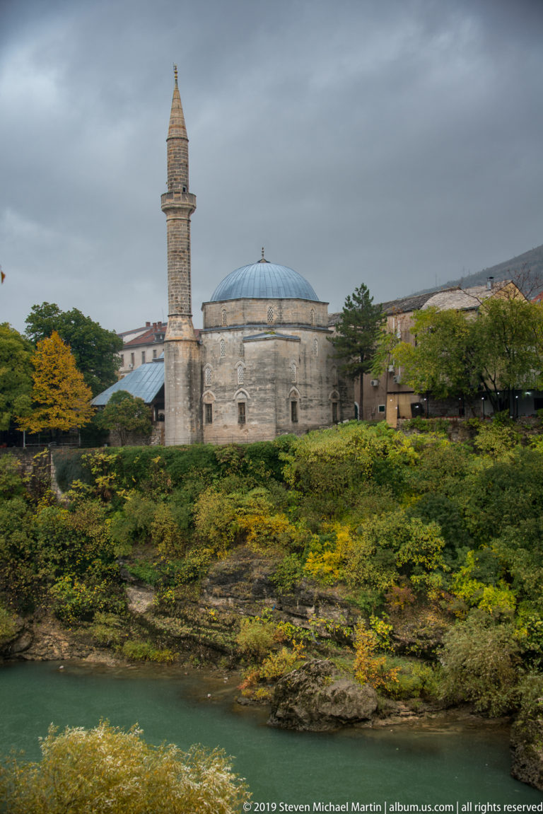 Stone mosque with a broad gray dome and tall minaret standing above the Neretva River amid autumn foliage.