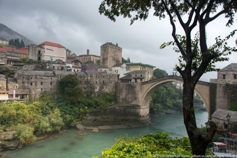 Stari Most over the Neretva River, framed by a large tree and surrounded by stone buildings and autumn vegetation.