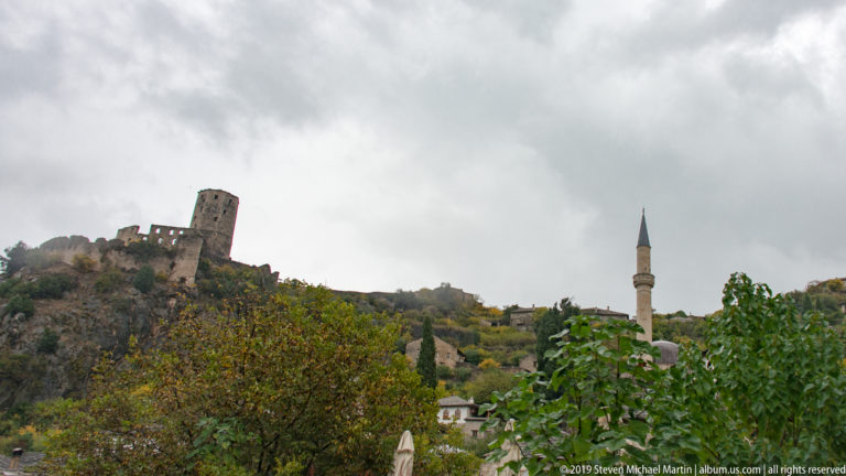 Počitelj's hilltop fortress, stone towers, and mosque minaret rising above autumn trees beneath a cloudy sky.