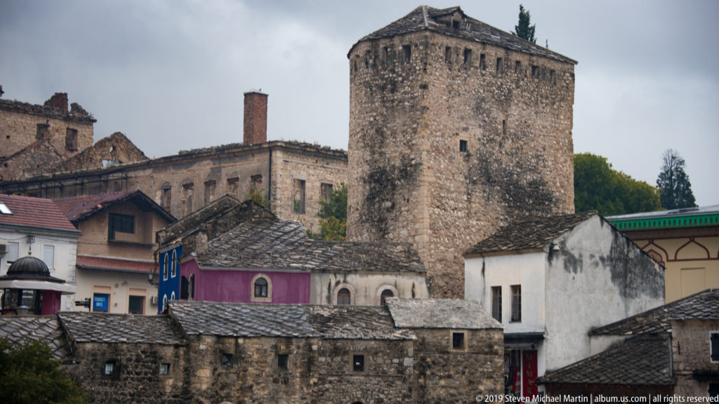 Large rectangular stone tower rising above closely packed historic stone houses and gray roofs in Mostar.