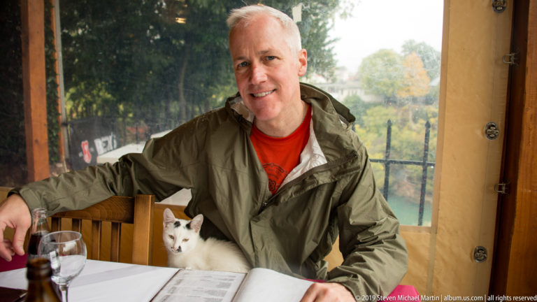 Man seated at a wooden restaurant table with a white cat beside him and a blurred riverside view behind.