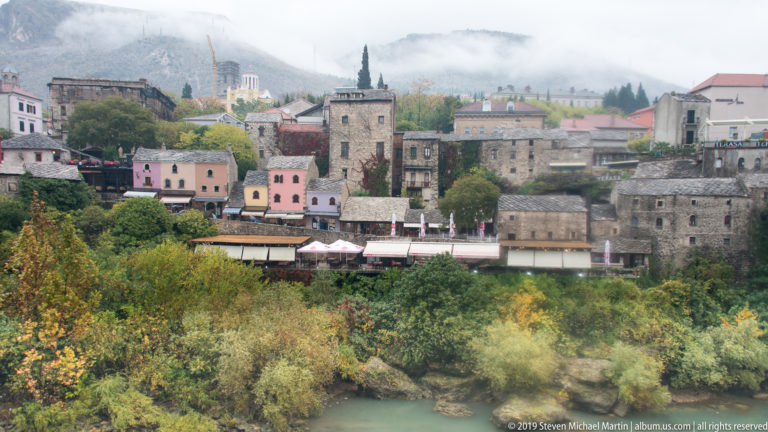 Cluster of stone and pastel buildings above the Neretva River, with trees in the foreground and cloudy mountains behind.