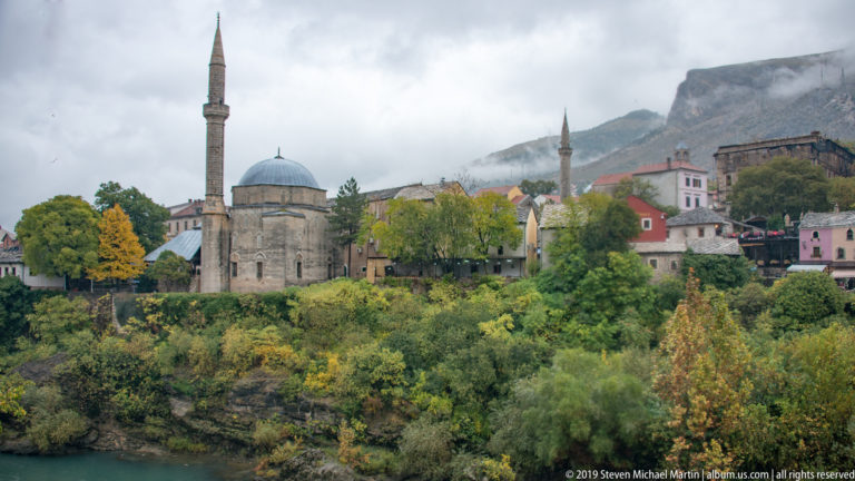 Domed stone mosque and tall minaret above the Neretva River, surrounded by autumn trees and misty mountains.