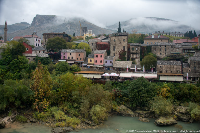 Stone and pastel buildings on a wooded bank of the Neretva River, backed by mist-covered hills.