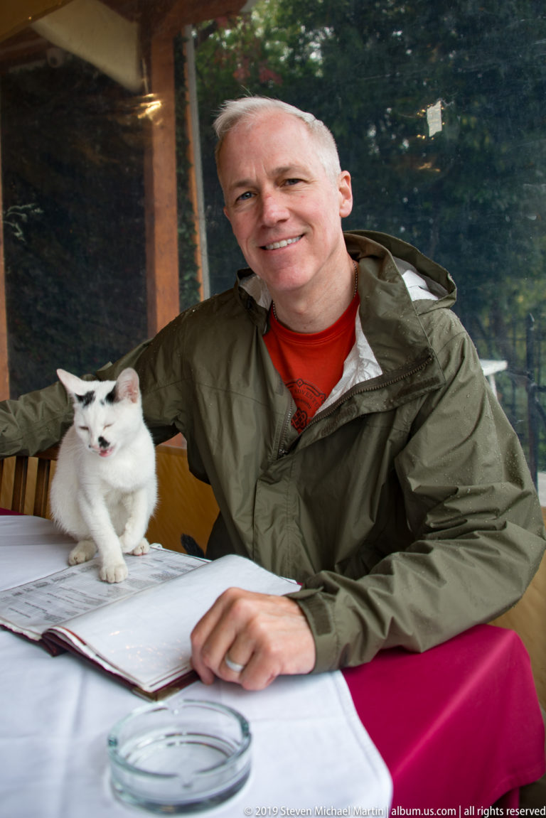 Man in a green jacket seated at a restaurant table beside a white cat, with a riverside view through the window.