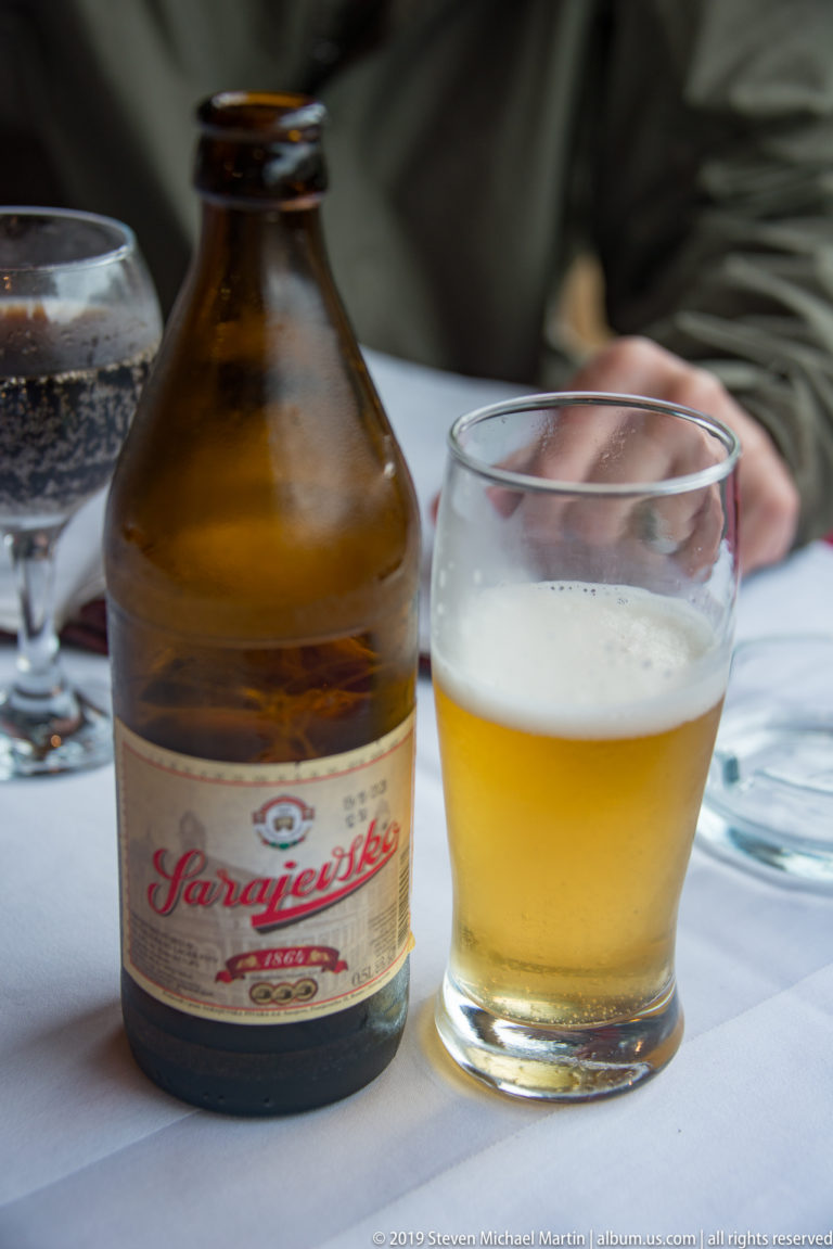Brown Sarajevsko beer bottle beside a small foamy glass of pale beer on a restaurant table.