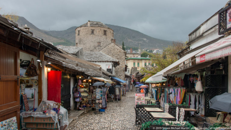 Narrow wet cobbled lane lined with souvenir shops and hanging textiles in Mostar's historic Kujundžiluk bazaar.