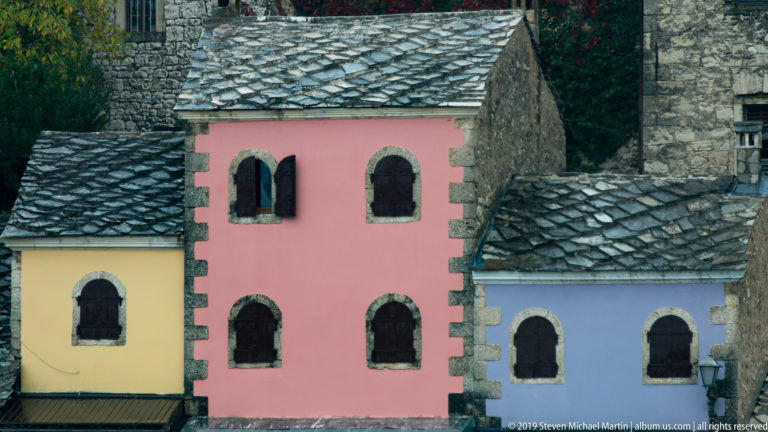 Yellow, pink, and blue historic façades with arched windows and dark stone-slab roofs in Mostar.