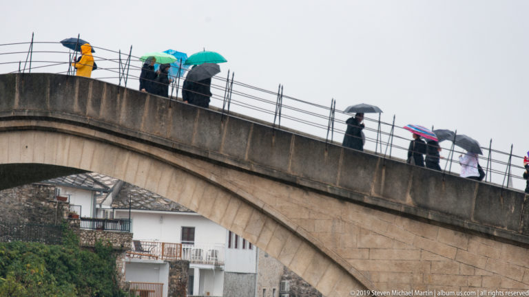 Pedestrians with umbrellas spaced along the rising stone parapet of Stari Most.