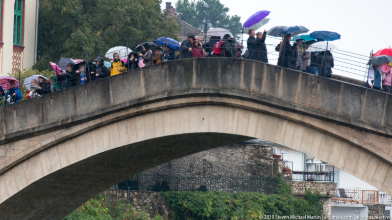 Crowd carrying colorful umbrellas across the steep crest of Stari Most in the rain.
