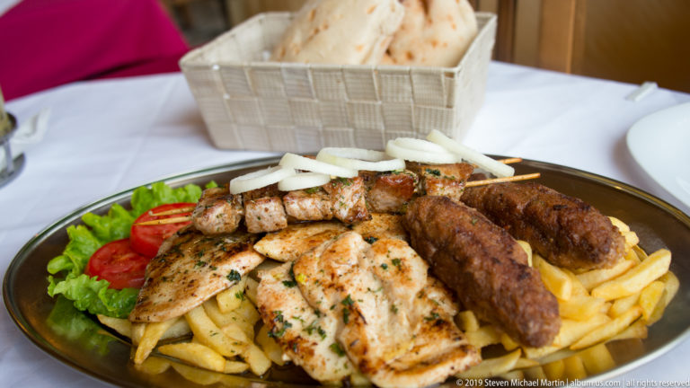 Plate of grilled chicken and minced-meat sausages with fries, onions, tomato, lettuce, and flatbread.