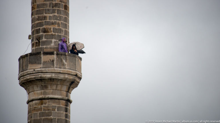 Two people, one with an umbrella, standing on the projecting balcony of a tall stone minaret.