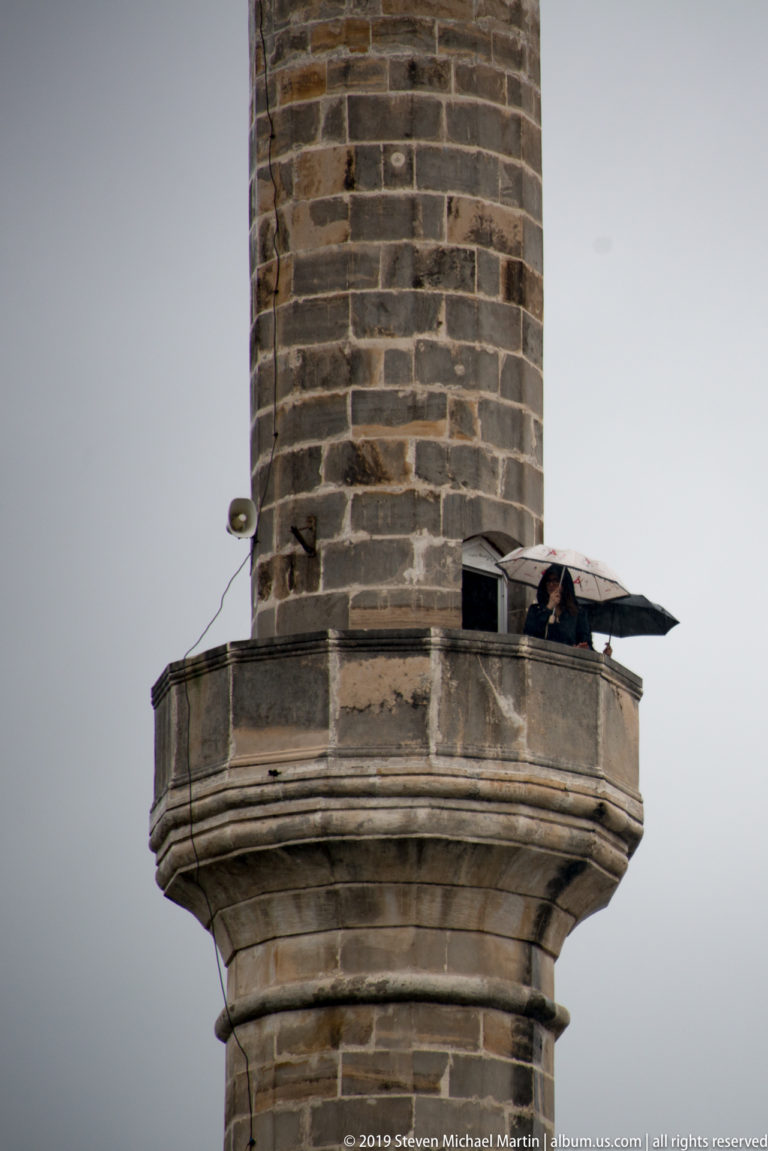Close view of the projecting balcony on a dark stone minaret against a gray sky.
